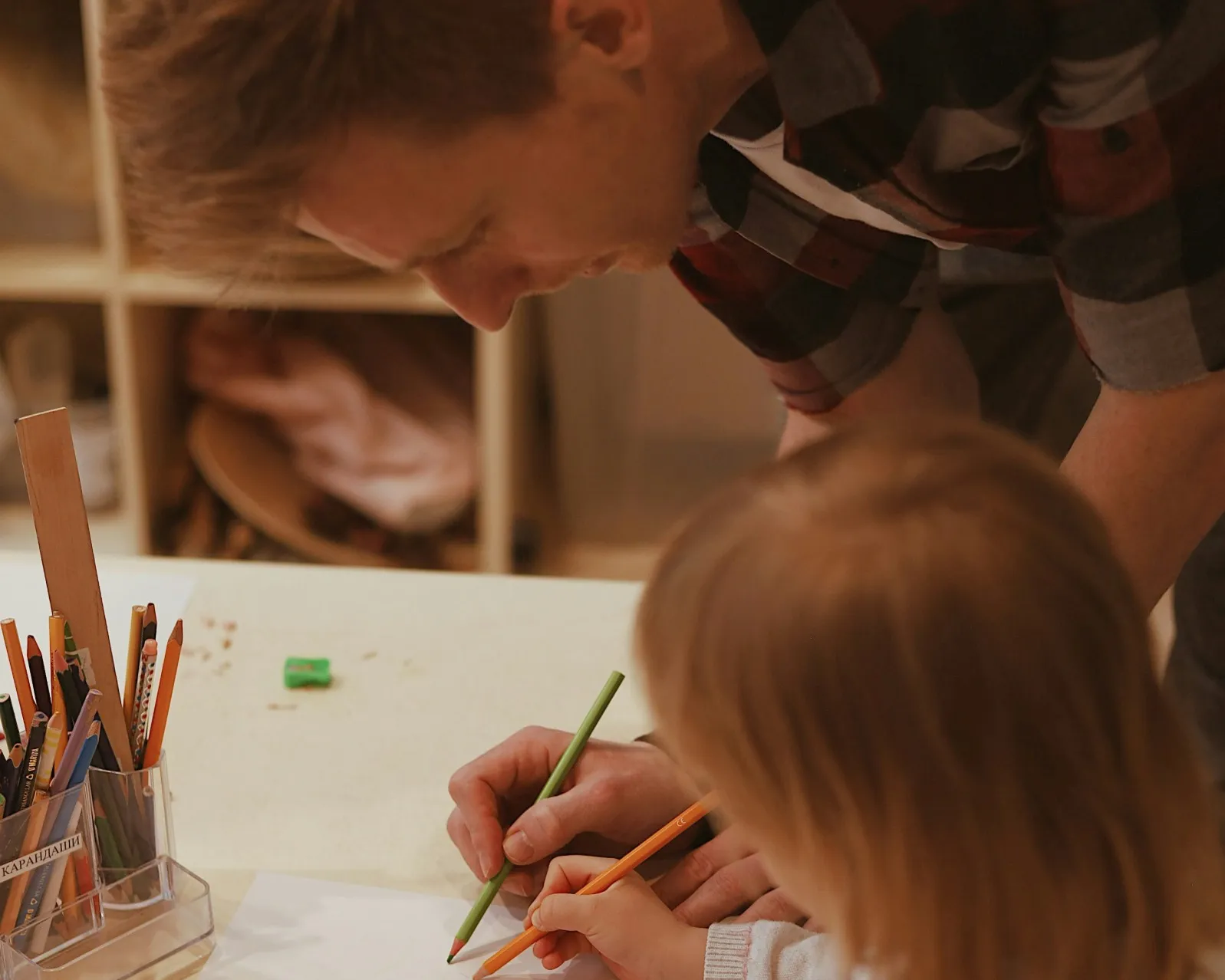 A man leaning in to guide a young child's pencil as they draw together at a craft table.